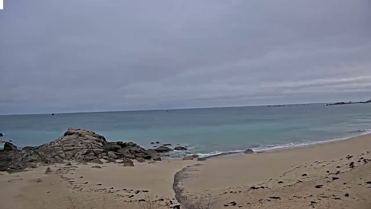 A sandy beach with dark rocks and a calm, light-blue ocean under an overcast sky.