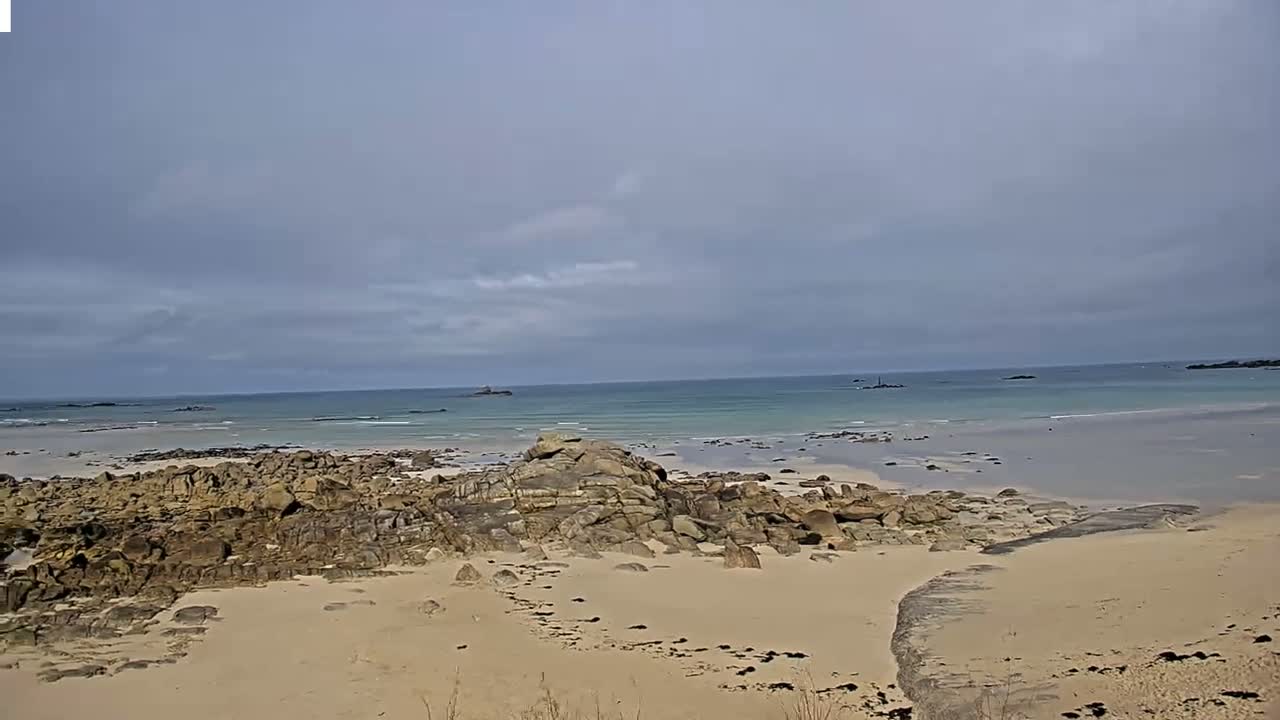 A sandy beach with dark rocks extends to a calm, light-blue ocean under a mostly cloudy sky.