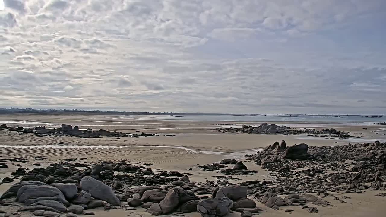 A sandy beach with dark rocks and small streams of water stretches toward a calm sea under a cloudy sky.