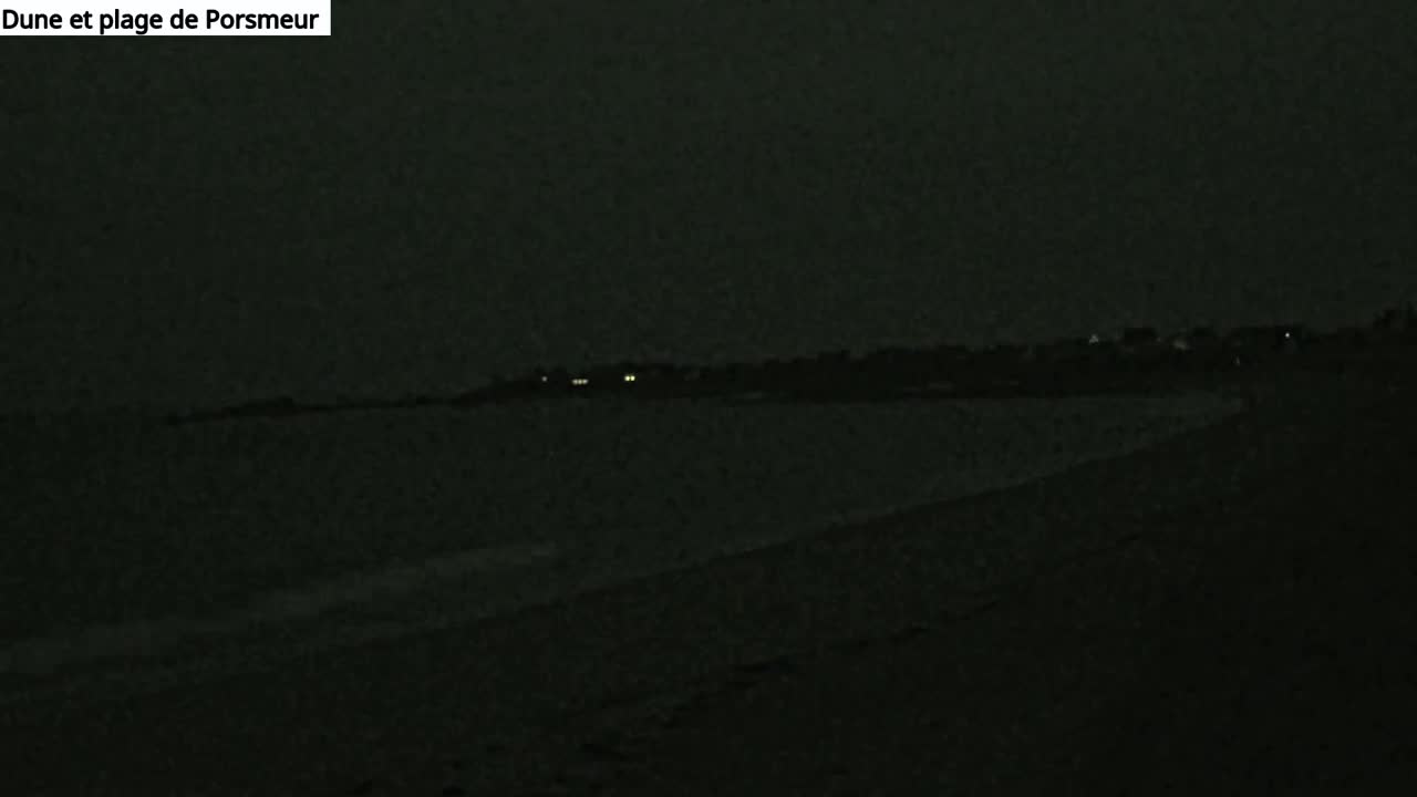 A dark, nighttime view of a beach and distant, dimly lit buildings under a cloudy sky.