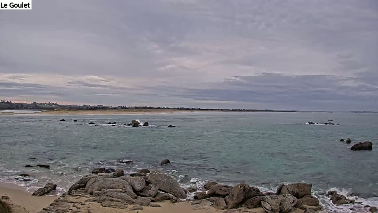 A rocky shoreline meets a calm, teal ocean under a mostly cloudy sky.