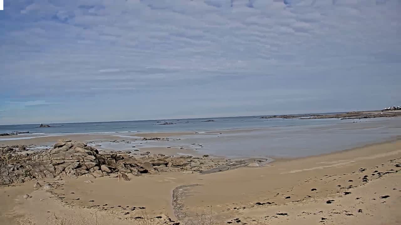 A sandy beach with rocky outcrops stretches towards a calm sea under a partly cloudy sky.
