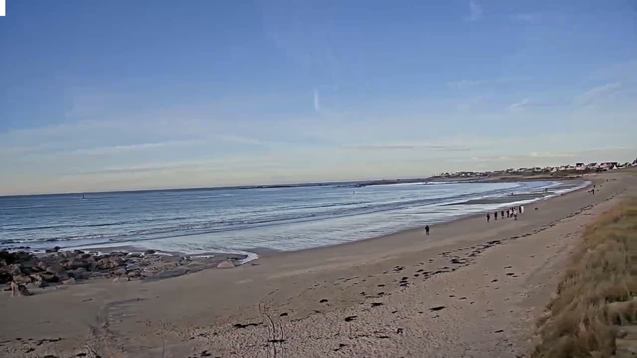 A sandy beach with gentle waves under a clear blue sky, showing a few people walking along the shoreline.