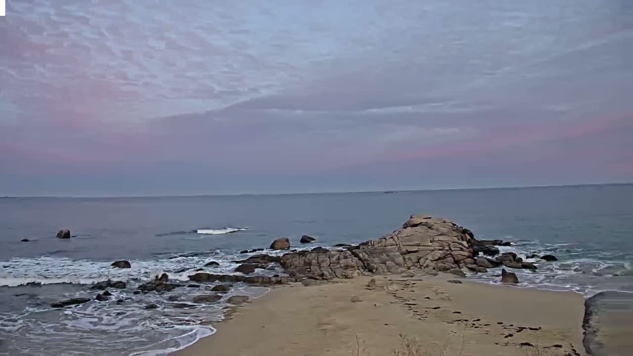 Under a pale pink and purple sky, gentle waves lap against a rocky shore and sandy beach.