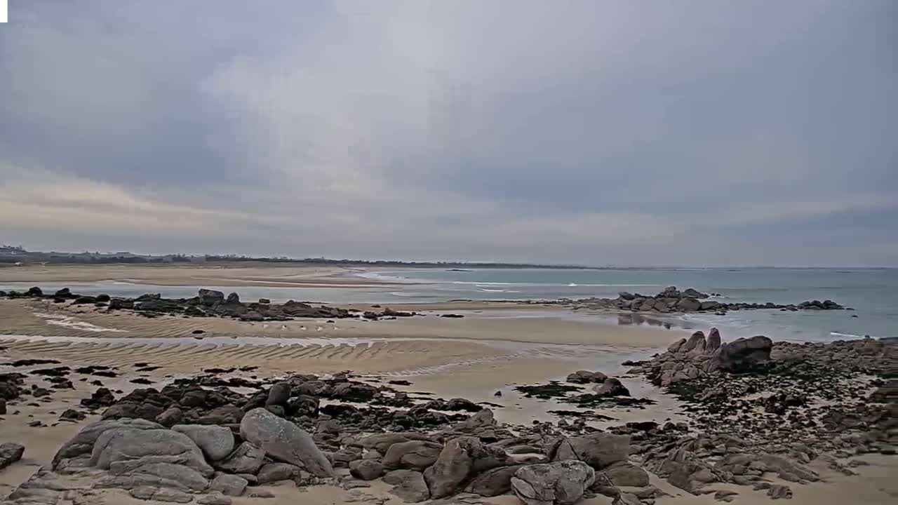 A sandy beach with dark rocks and small waves under an overcast sky stretches to a calm ocean horizon.