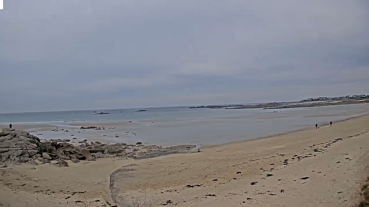 A sandy beach stretches towards a calm, light-grey sea under an overcast sky, with a few people walking along the shore and rocky outcrops near the water's edge.