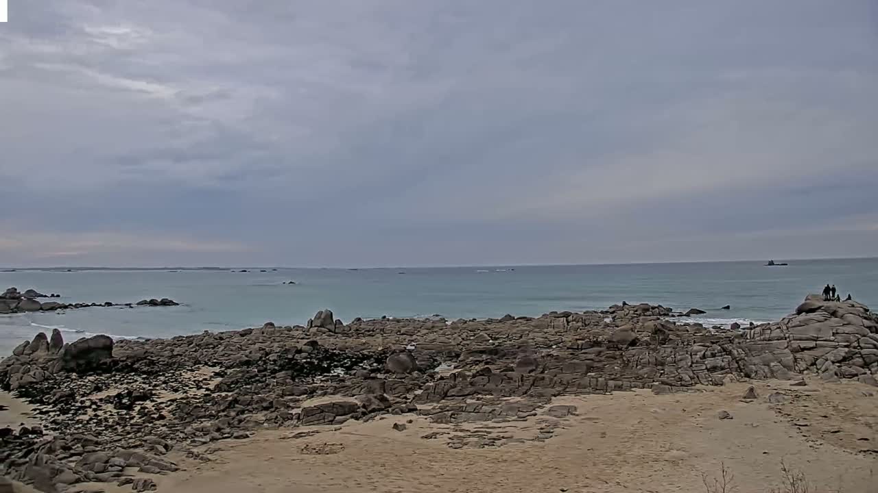 A rocky beach stretches to a calm, light teal ocean under a cloudy sky.