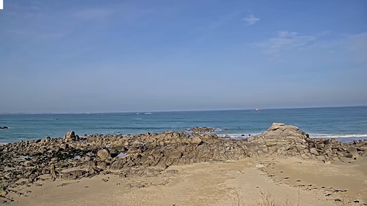 A sandy beach with dark rocks and a calm, light blue ocean under a mostly clear sky.