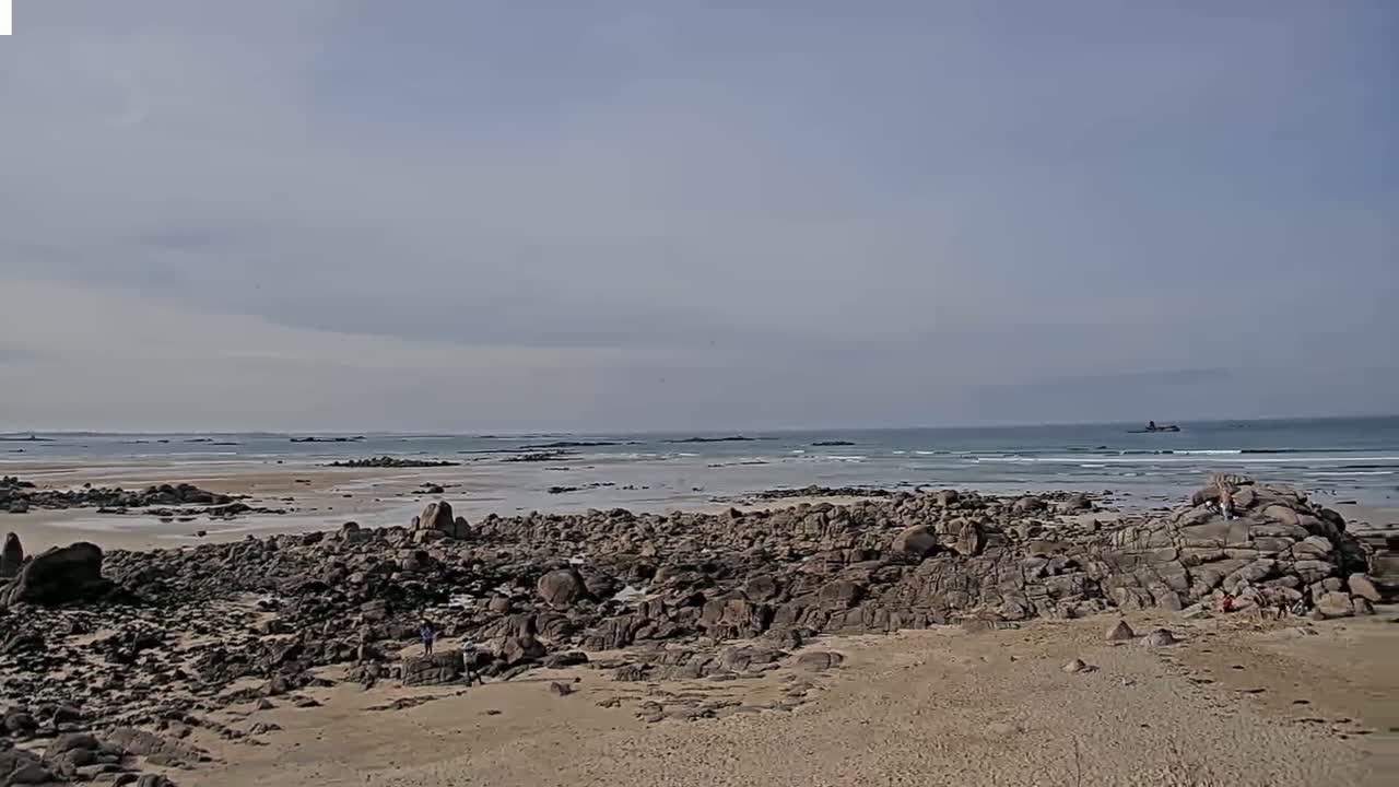 A sandy beach with dark-colored rocks and a calm ocean under a mostly cloudy sky.
