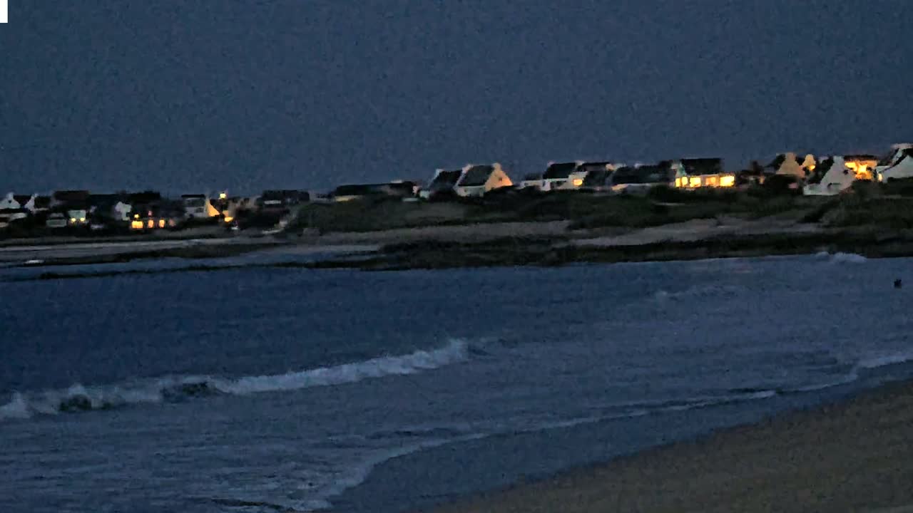 Small waves lap a dark sandy shore at dusk, with a line of houses with lit windows visible on a distant hill.