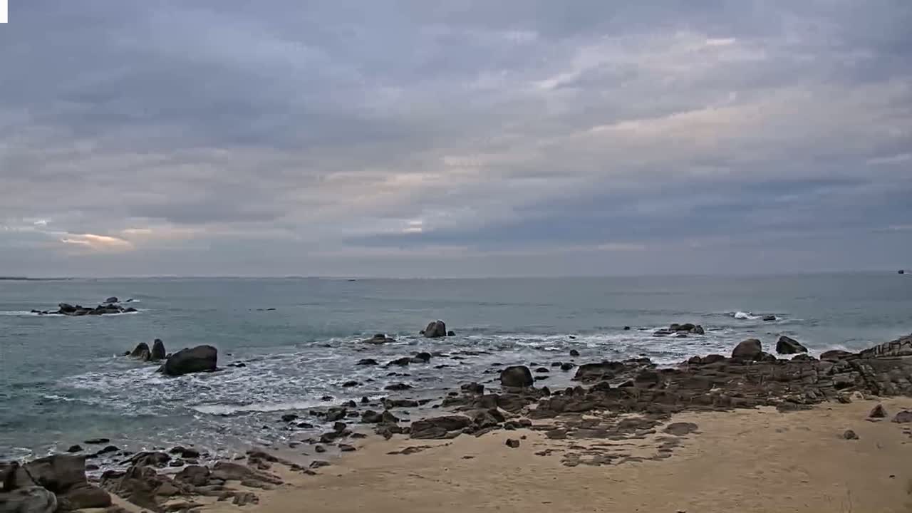 A rocky shoreline with a sandy beach stretches out to a calm, gray sea under a cloudy sky.