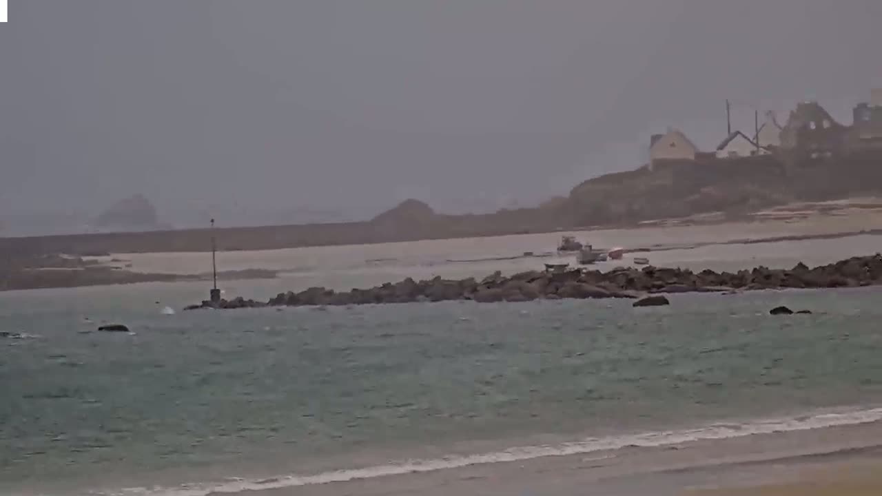 A rocky shoreline with a few small boats is visible in the distance under an overcast sky, with choppy water in the foreground.