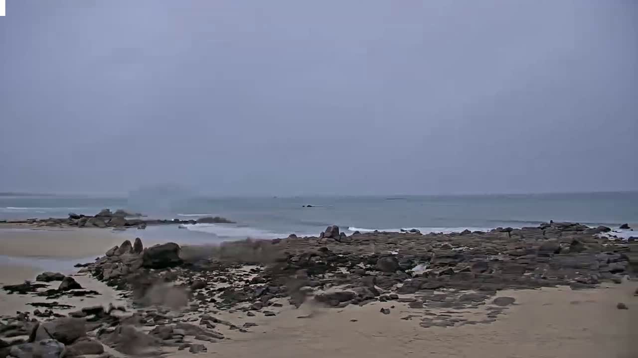 A sandy beach with dark rocks and a calm, overcast ocean stretches to a hazy horizon.