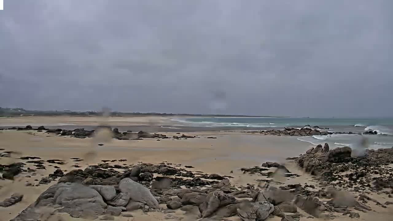 A sandy beach with dark rocks and small waves under an overcast sky, with rain visible on the camera lens.
