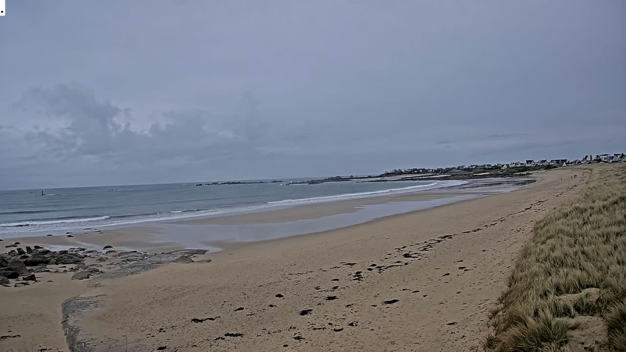 A sandy beach stretches to a calm ocean under a cloudy sky, with a small town visible in the distance.
