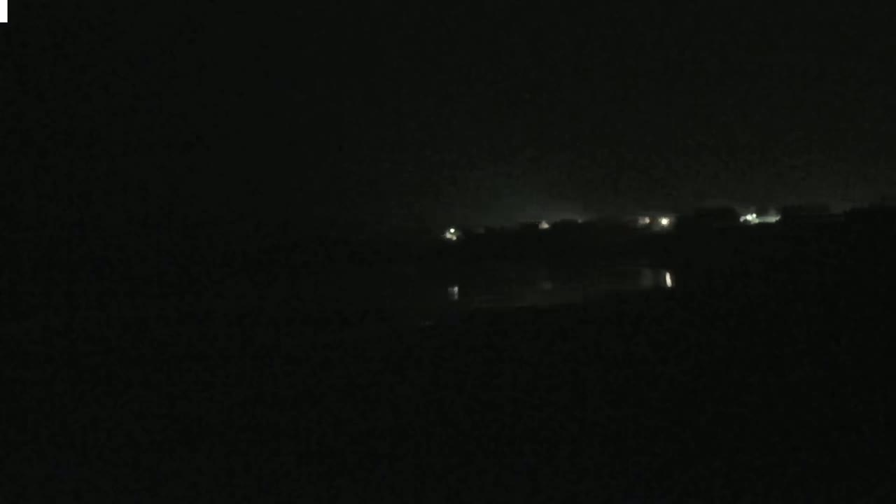 A dark, nighttime scene shows faintly lit buildings in the distance across a body of still water.