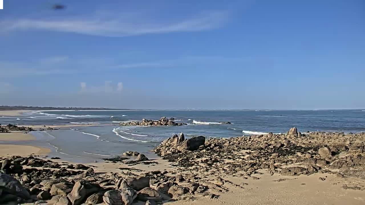 A sandy beach with dark rocks and small waves under a mostly clear blue sky.