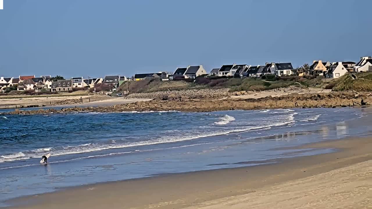A sandy beach with small waves laps against a rocky shore, in front of a line of houses under a clear blue sky.