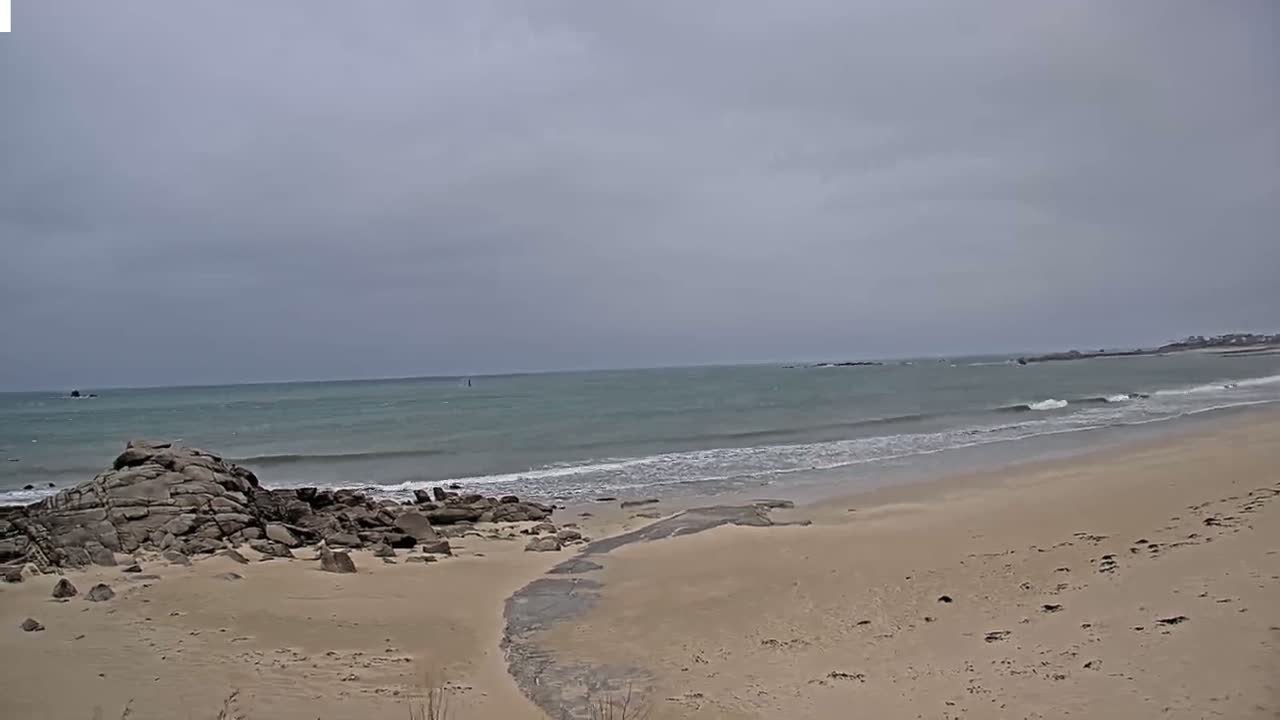 A sandy beach with dark rocks near the water's edge meets a calm, grey ocean under an overcast sky.