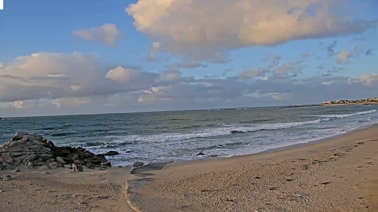 A sandy beach with rocky outcrops meets a gently wavy ocean under a partly cloudy sky.