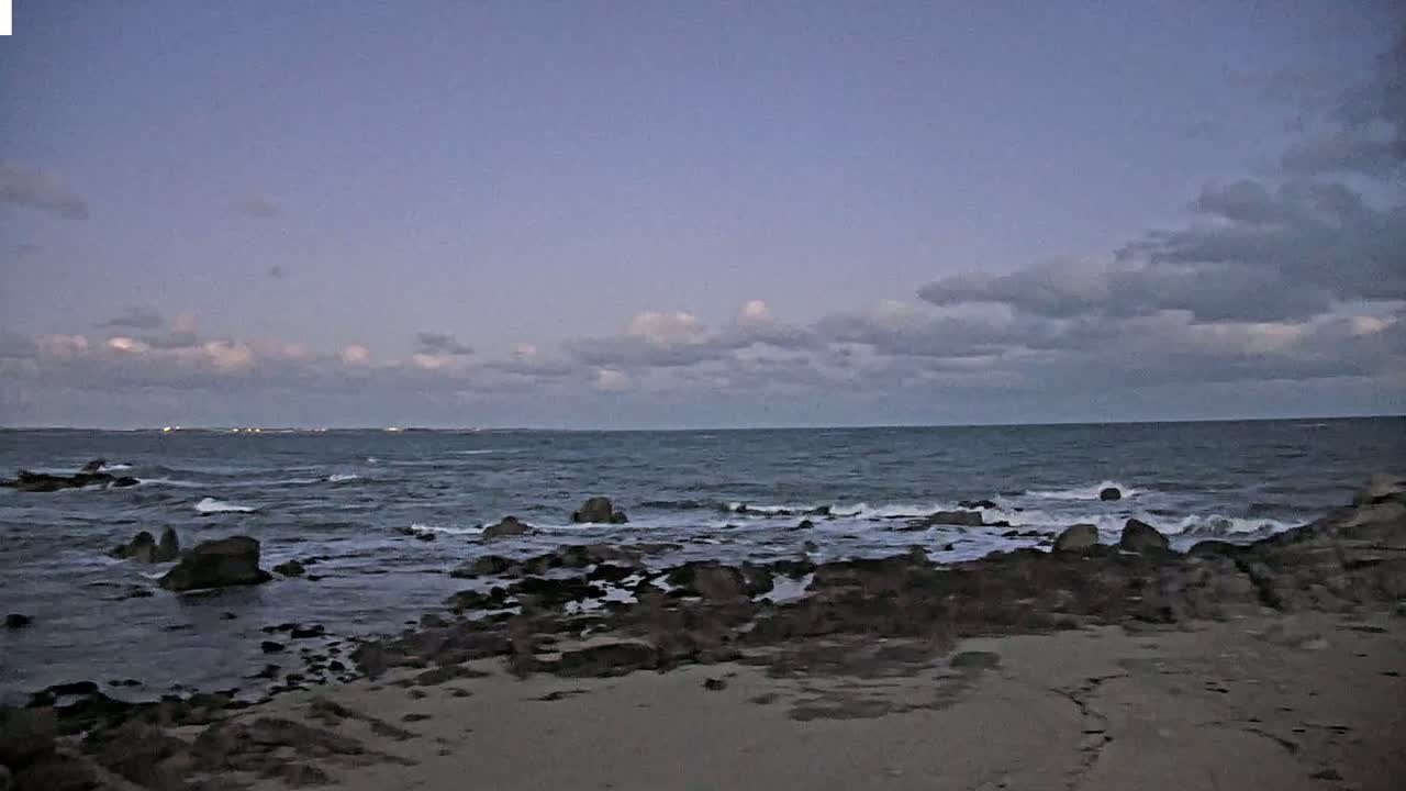 A rocky shoreline with gentle waves under a mostly cloudy, dusky sky.