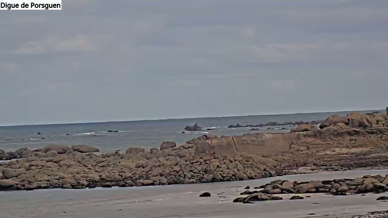 A rocky coastline meets a calm, gray sea under a mostly cloudy sky.