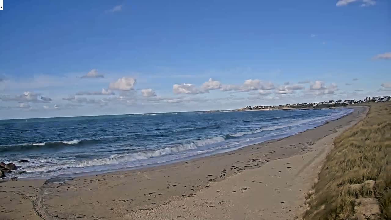 A sandy beach curves gently around a bay with small waves lapping the shore under a mostly sunny sky with scattered clouds.