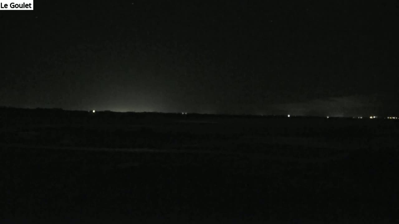 A dark, nighttime landscape shows a distant horizon line illuminated by faint, scattered lights under a clear, starlit sky.