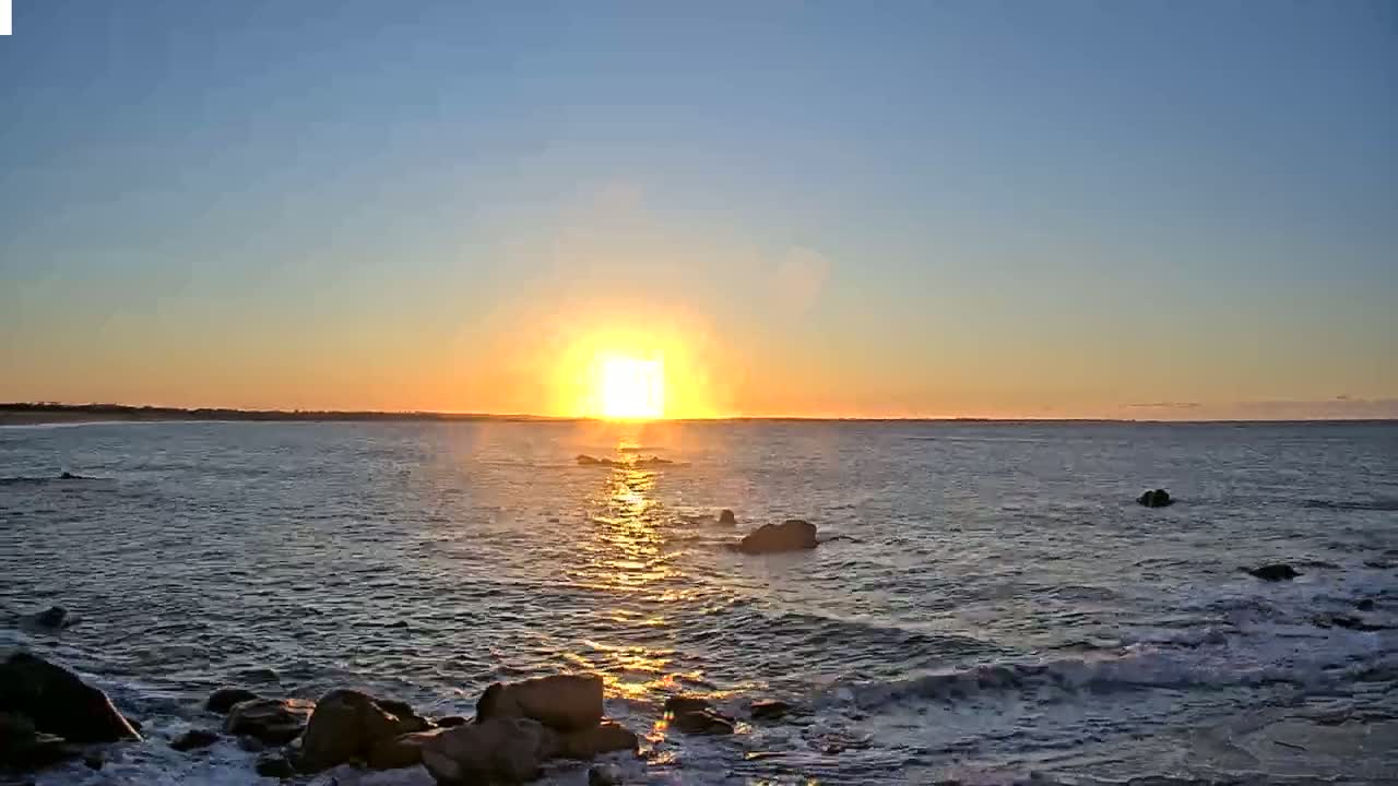 A bright, orange sunset over a calm ocean with dark rocks in the foreground.
