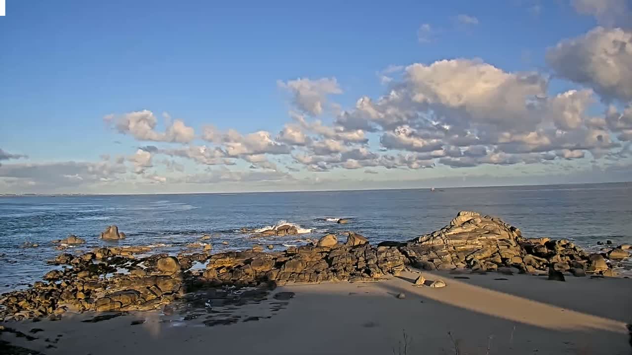 A sandy beach with dark rocks extends into a calm ocean under a mostly sunny sky with scattered clouds.
