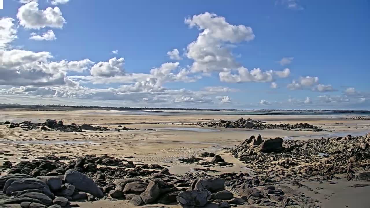 A mostly sunny day reveals a sandy beach with dark rocks in the foreground, extending to a calm body of water under a partly cloudy sky.
