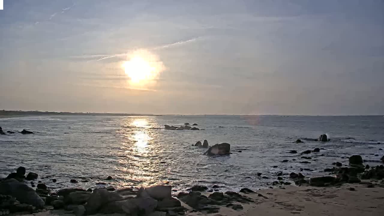 A calm ocean with dark rocks along the shoreline reflects the bright sun during a partly cloudy sunset.