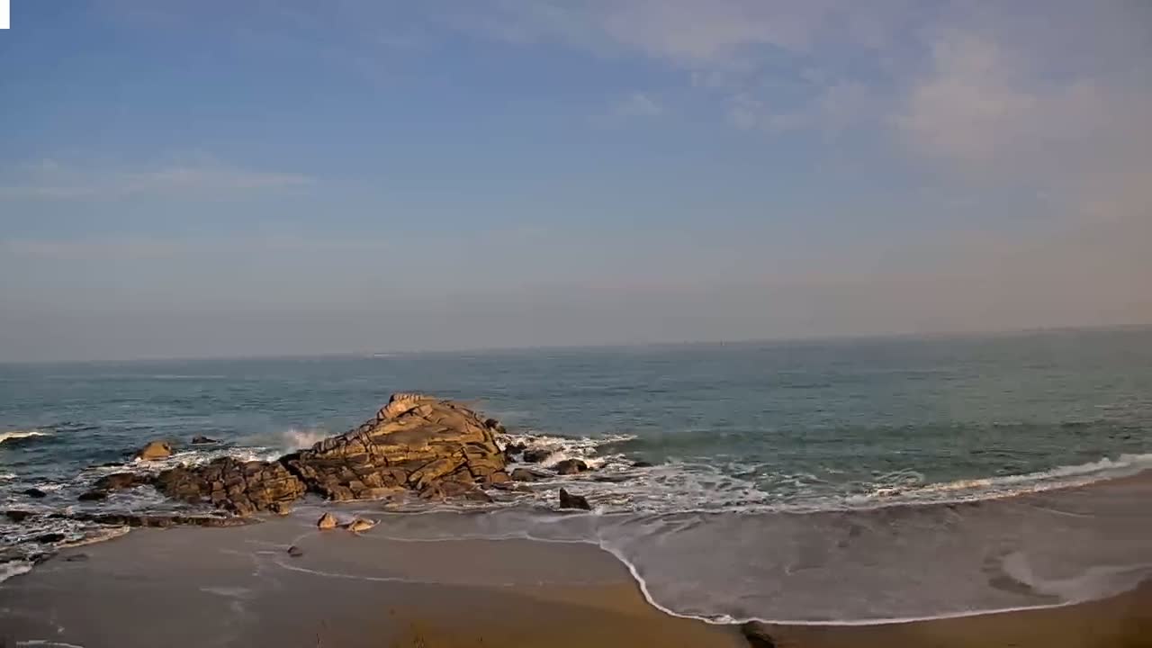 Under a mostly clear sky, gentle waves lap a sandy beach near a rocky outcrop.