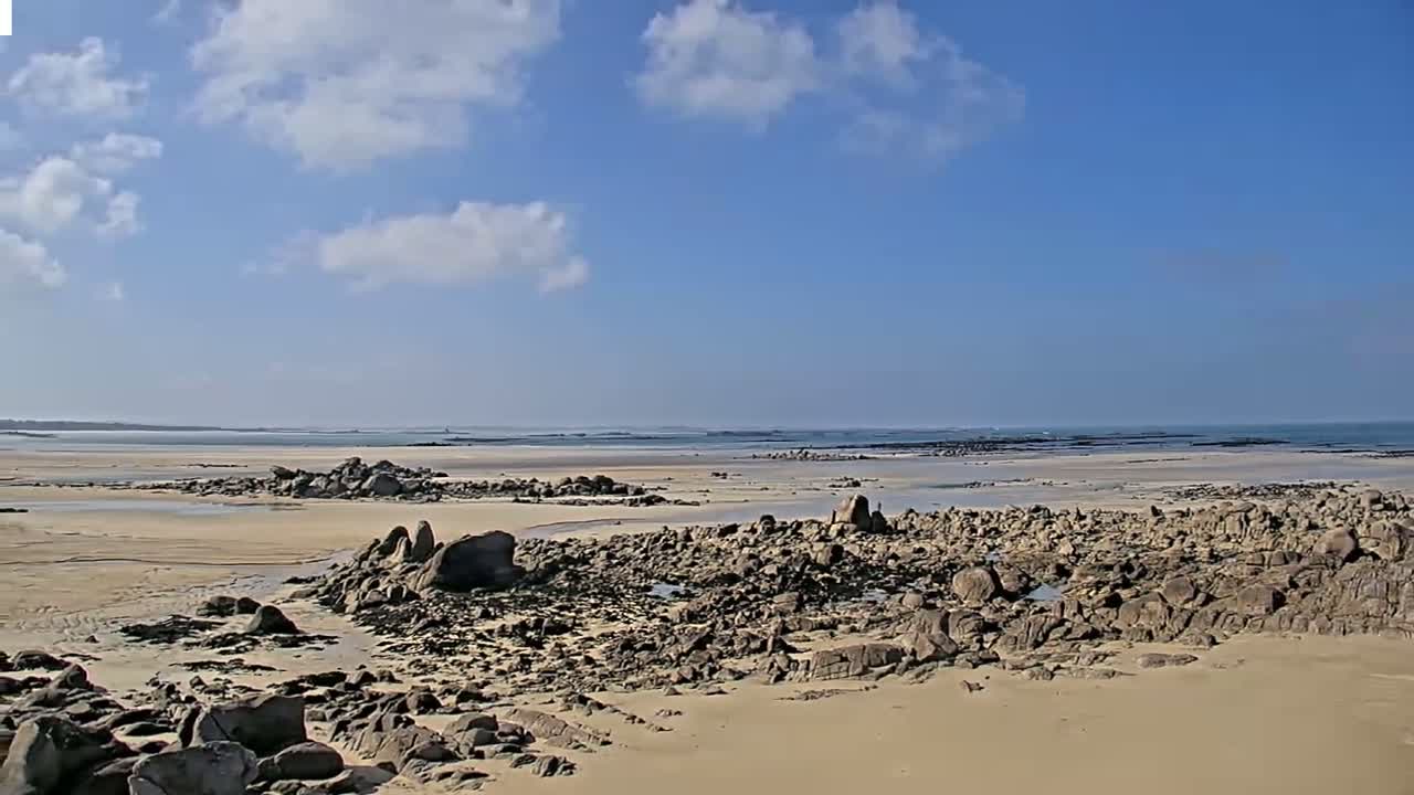 A sandy beach with dark rocks and shallow pools stretches to a calm ocean under a mostly sunny sky with scattered clouds.