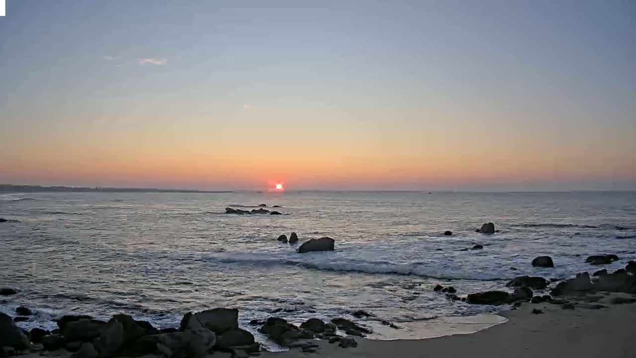 The setting sun casts a warm glow over a calm ocean, where gentle waves lap against a rocky shoreline.