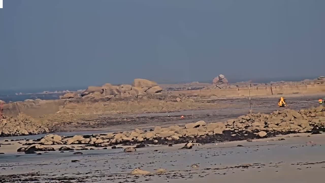A rocky, sandy beach stretches to a calm ocean under a clear, hazy sky, with some construction equipment visible in the mid-ground.