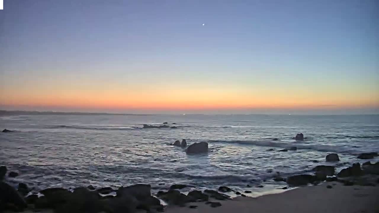 A calm ocean at sunset with dark rocks on a sandy shore under a clear, twilight sky.