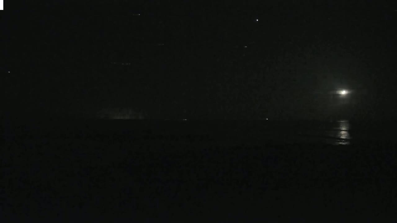 A dark, moonlit night scene shows a body of water reflecting the moon's light, with a faint horizon line and a few distant lights visible.