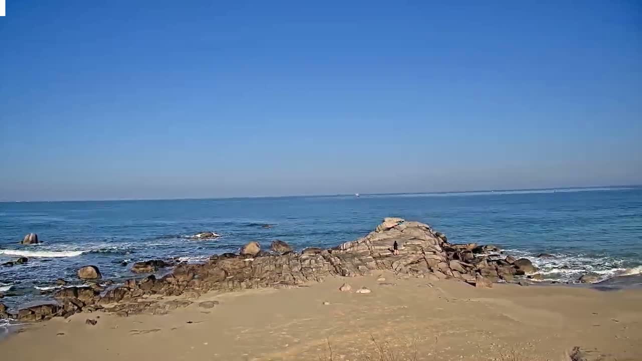 A sandy beach with dark rocks extends into a calm, blue ocean under a clear blue sky.