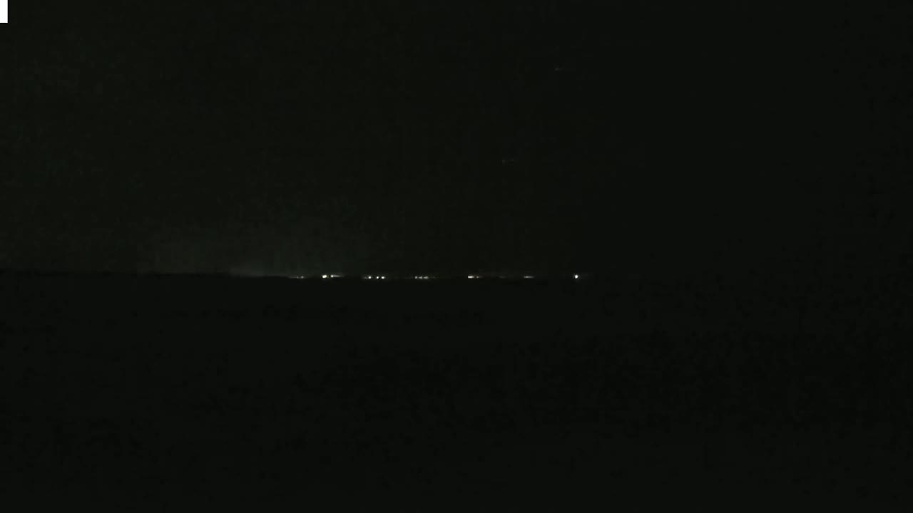 A dark, nighttime landscape shows a faint line of distant lights on the horizon under a mostly clear sky.