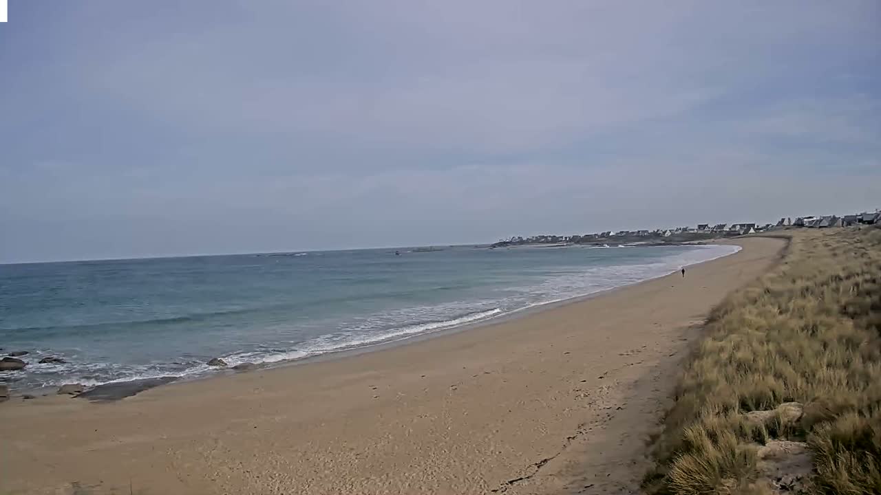 A sandy beach with small waves under a cloudy sky curves gently towards a distant line of houses.