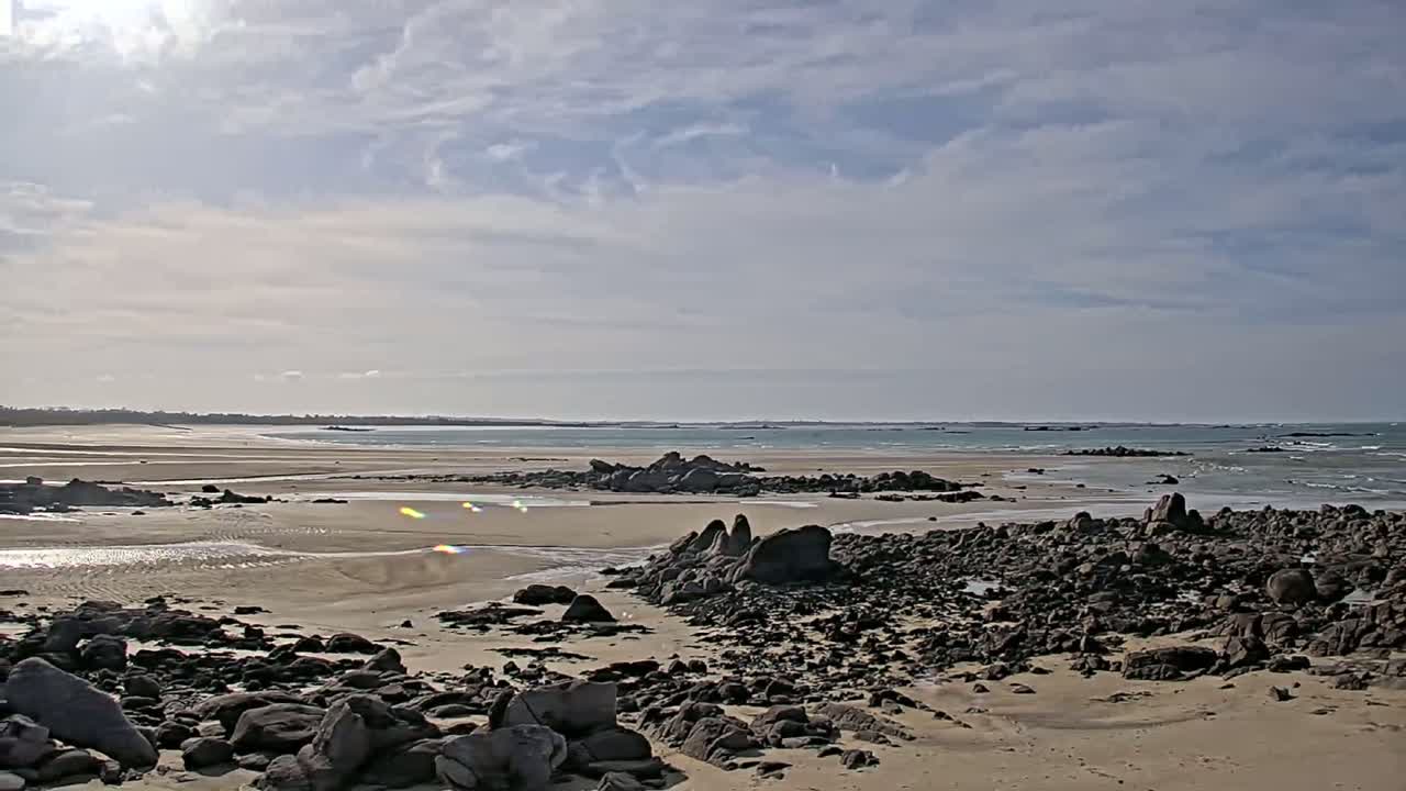 A sandy beach strewn with dark rocks extends to a calm, light-blue ocean under a partly cloudy sky.