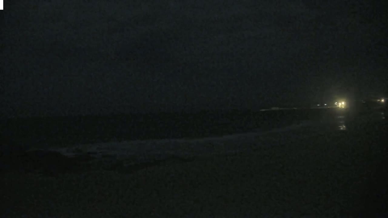 A dark nighttime beach scene shows a dark ocean with faint waves under a cloudy sky, and distant lights are visible on the right.