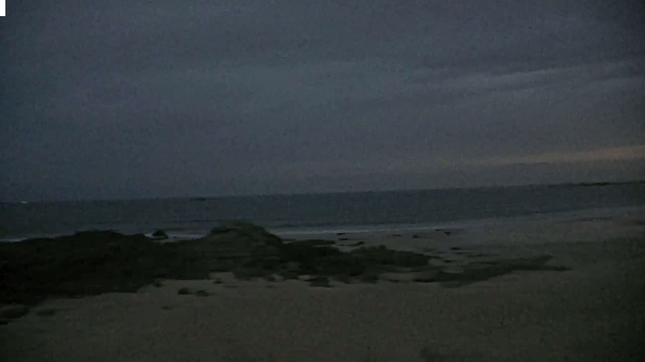 Dark, overcast skies loom over a calm, grey ocean meeting a sandy beach with dark rocks in the foreground.