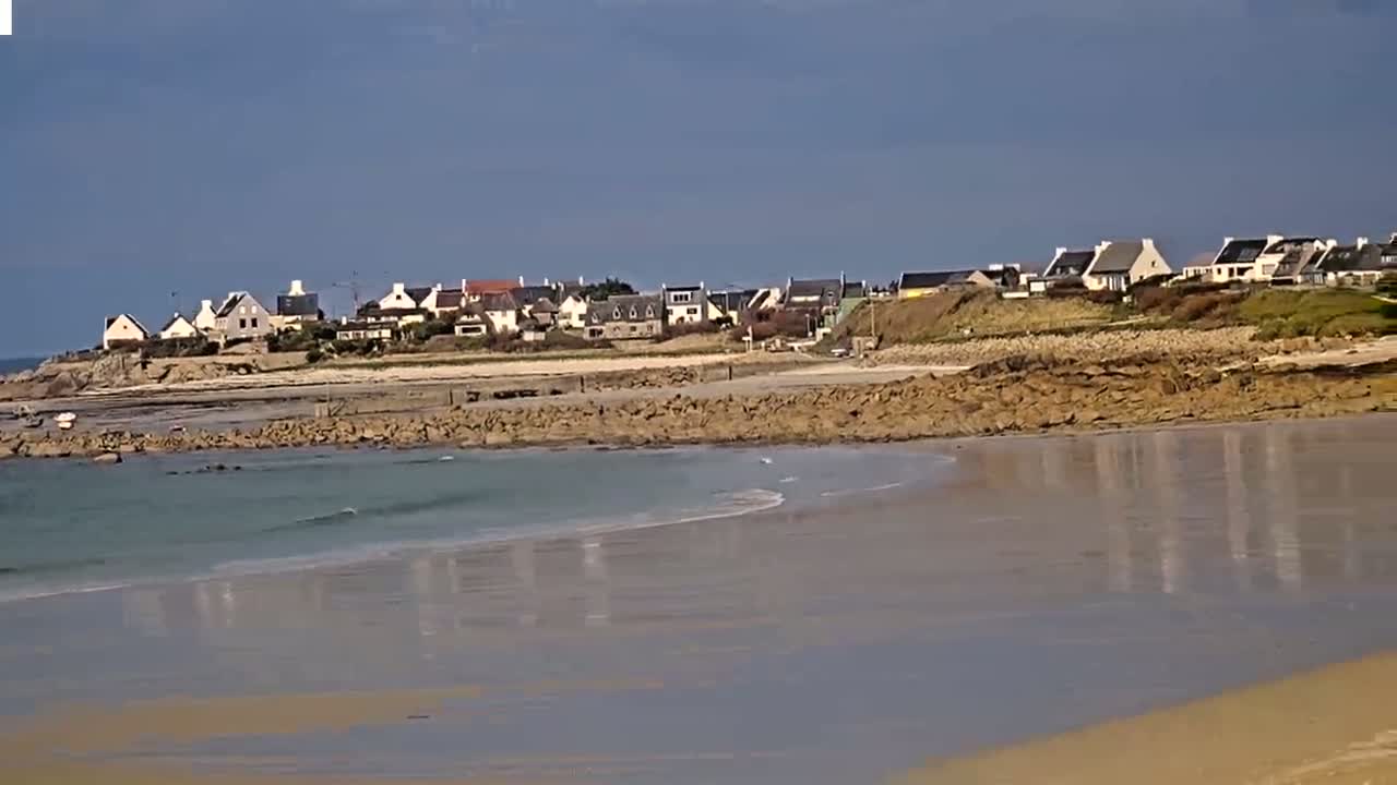 A sandy beach with calm, light-blue water reflects a mostly clear sky, and a line of houses sits atop a rocky shoreline in the background.