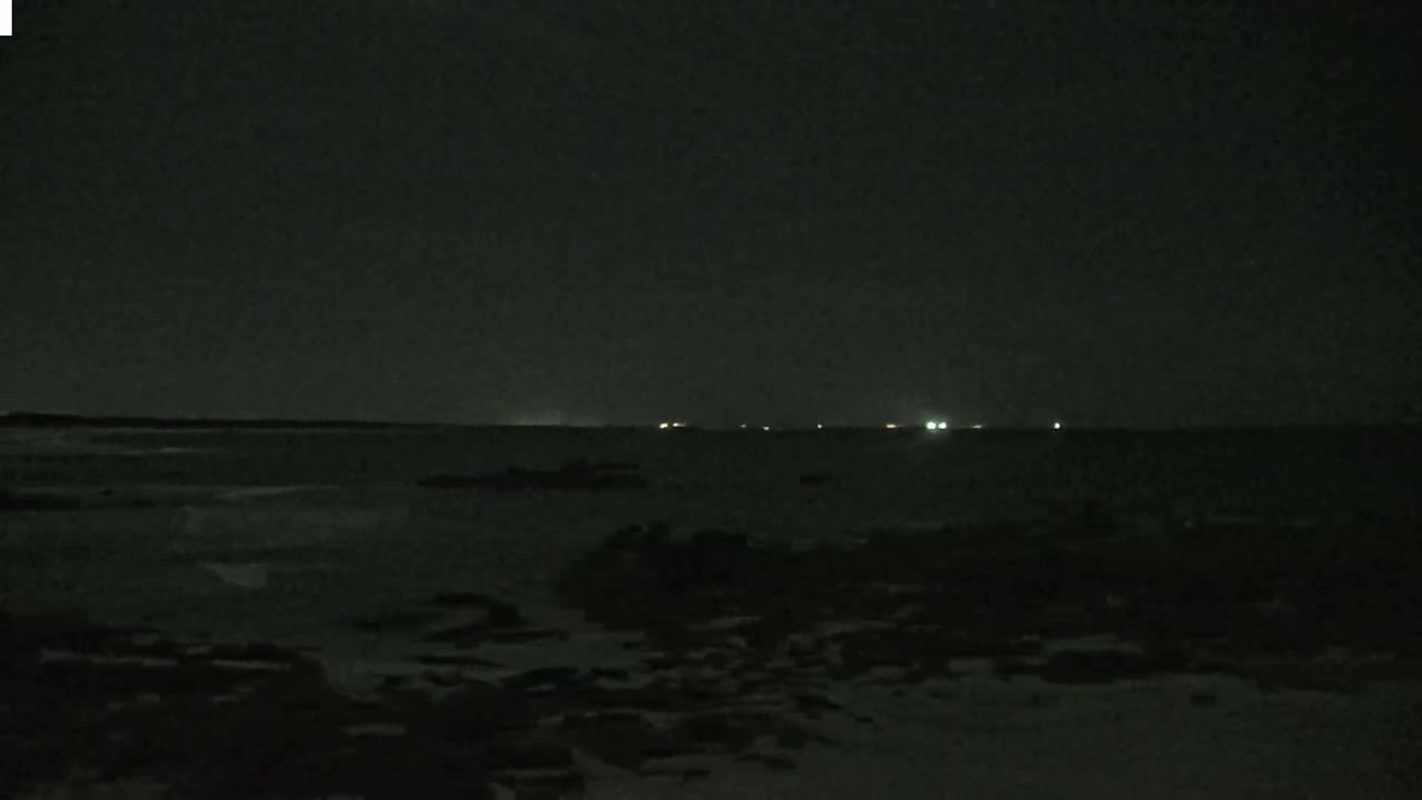 A dark, nighttime scene shows a rocky shoreline with a calm body of water extending to a distant horizon where faint lights are visible.