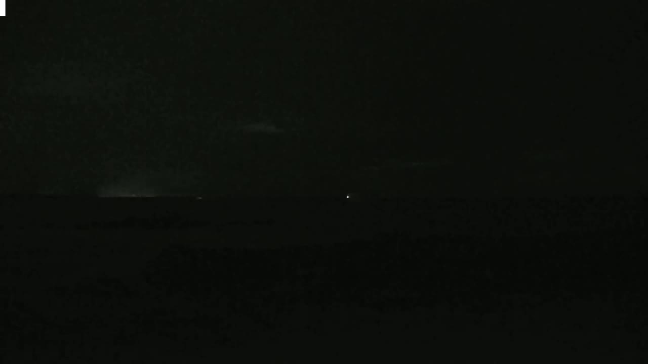 A dark, nighttime landscape is shown with a faint, distant light source and a slightly brighter horizon line suggesting a possible thunderstorm in the distance.
