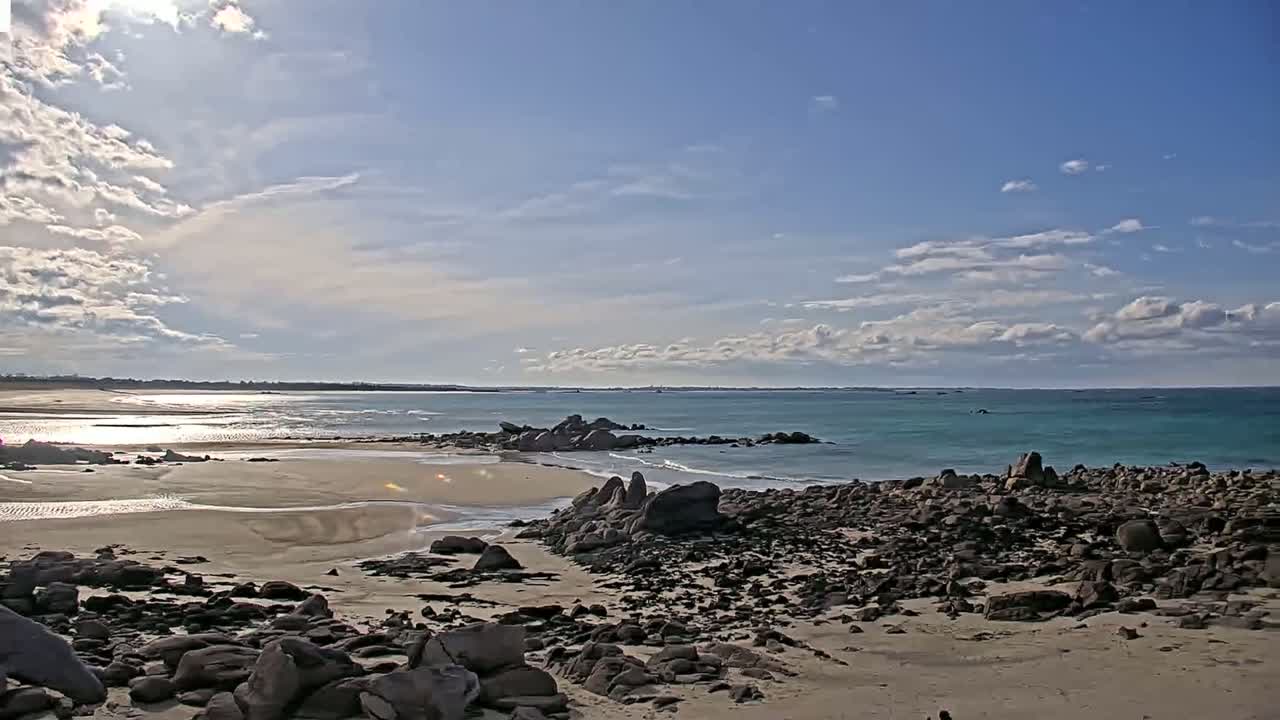 A sandy beach with dark rocks stretches to a calm, teal ocean under a partly cloudy sky.