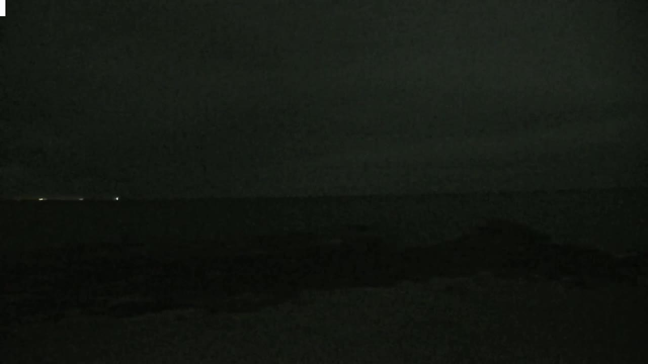 A dark, nighttime scene shows a body of water with a few faint lights visible on the horizon under a mostly clear, dark sky.