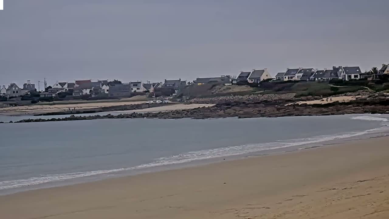 A sandy beach with gentle waves laps against the shore, in front of a line of houses nestled on a rocky coastline under an overcast sky.